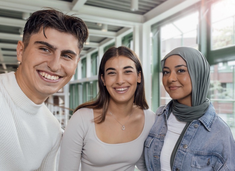 Three young colleagues smiling together in a bright office hallway.