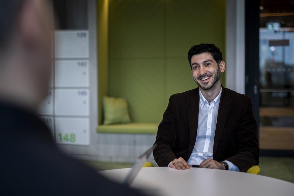 Man in a suit jacket smiling while sitting in a modern office with green seating and lockers in the background.