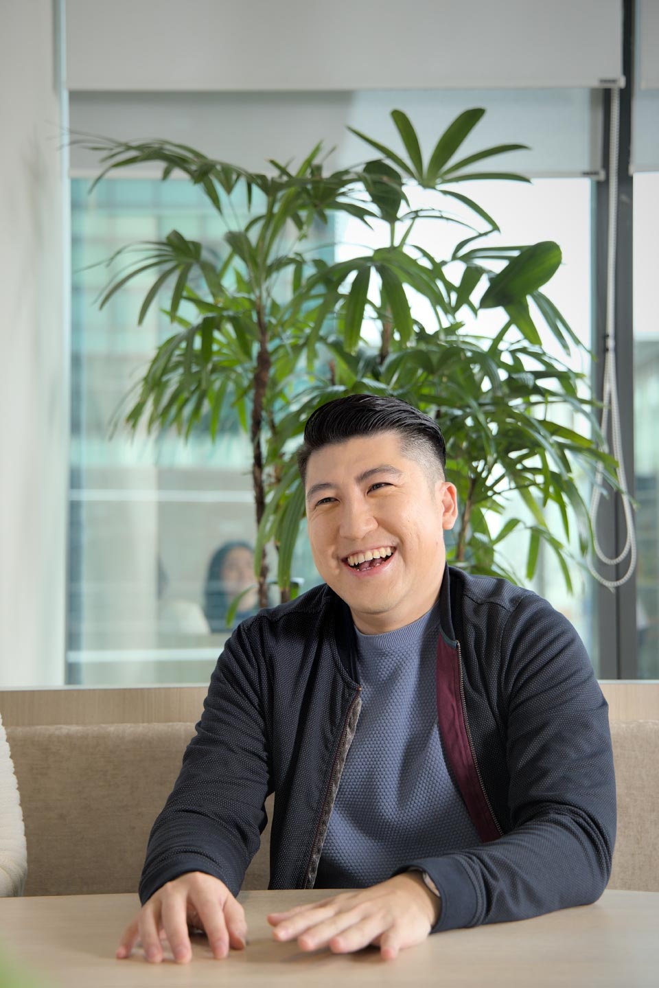 Man smiling warmly while sitting indoors with a plant in the background, creating a bright and friendly atmosphere.