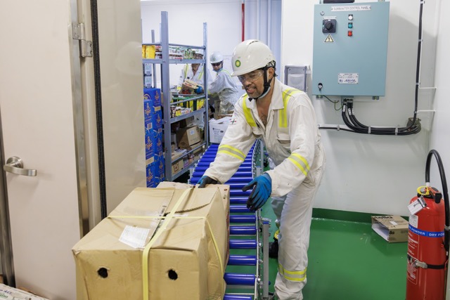 Worker in protective gear moving a large box on a conveyor inside an industrial facility.