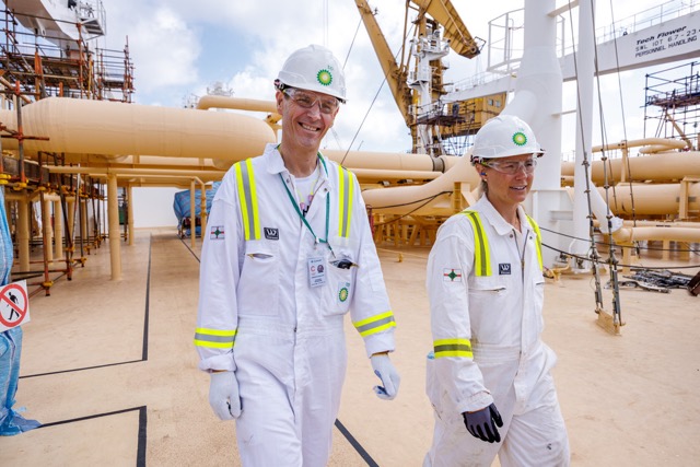 Two bp workers in white protective uniforms and helmets walking at an industrial site.