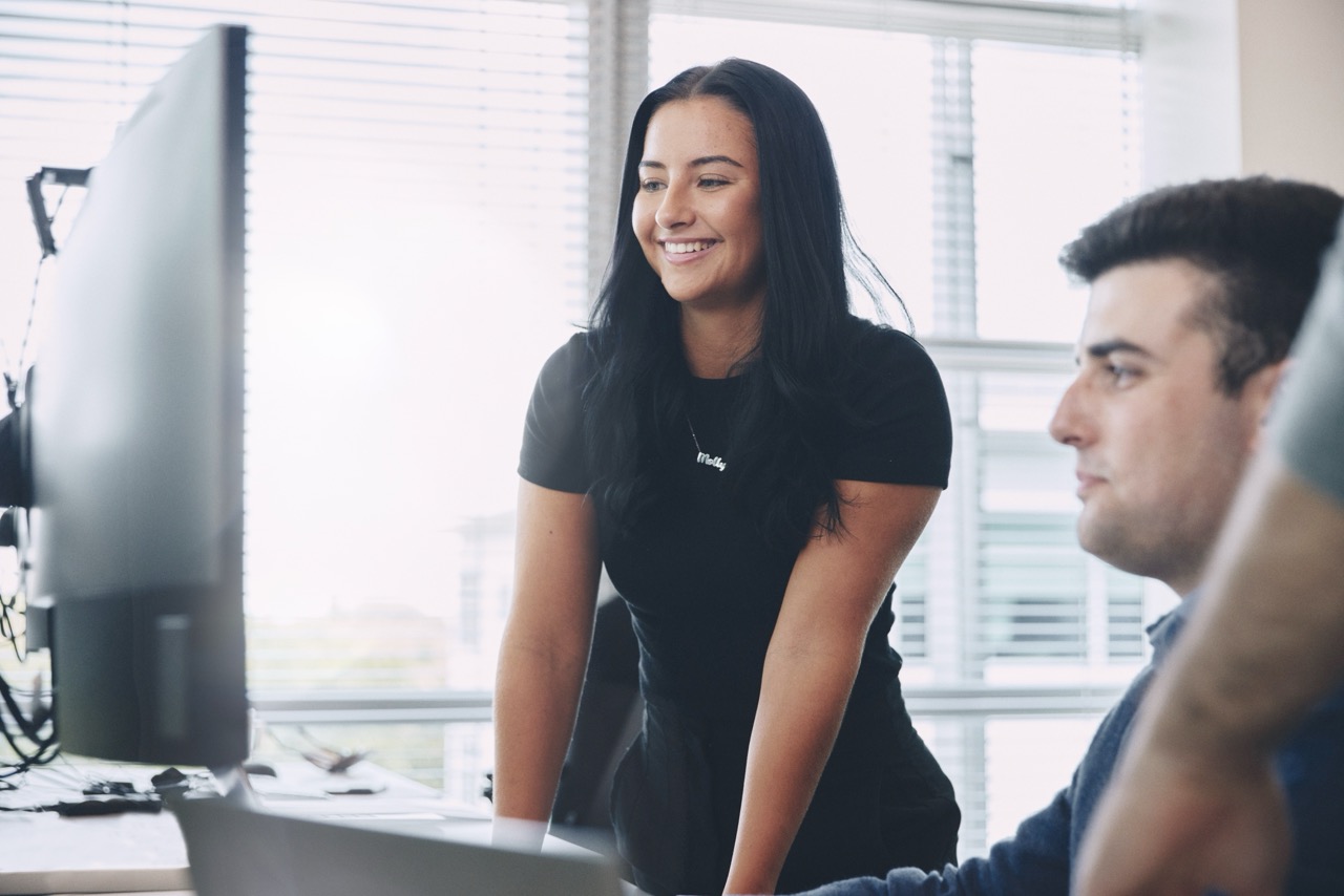 Smiling colleagues collaborating at computer workstations in a bright office.