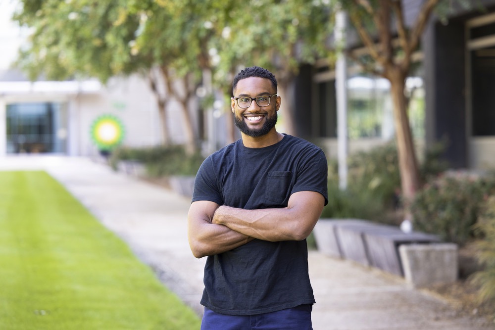 Man standing outdoors with arms crossed, smiling at the camera. He is wearing glasses and a black t-shirt, with an office building and greenery in the background.
