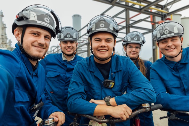 Group of five industrial workers in blue protective uniforms and helmets smiling at an outdoor worksite.