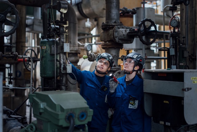Two industrial workers in blue uniforms and helmets inspecting equipment inside a facility.