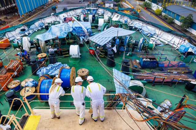 Three workers in white protective uniforms and helmets standing on a deck overlooking industrial equipment and cables.