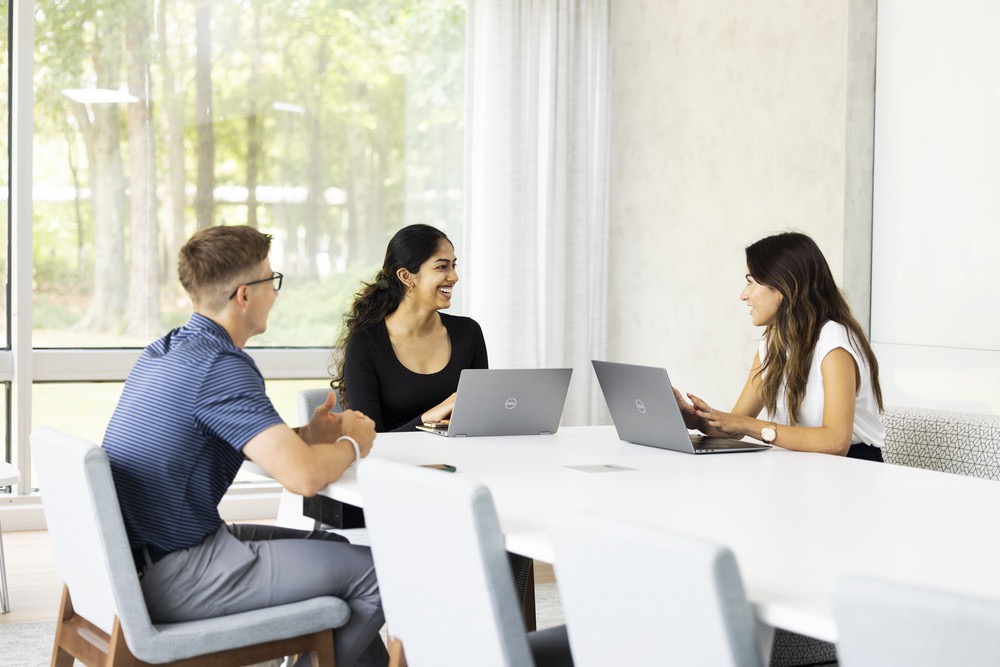Three colleagues sitting at a conference table with laptops, engaged in discussion.