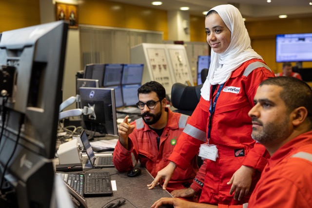 Three bp employees in red safety uniforms working together at computer monitors in a control room.