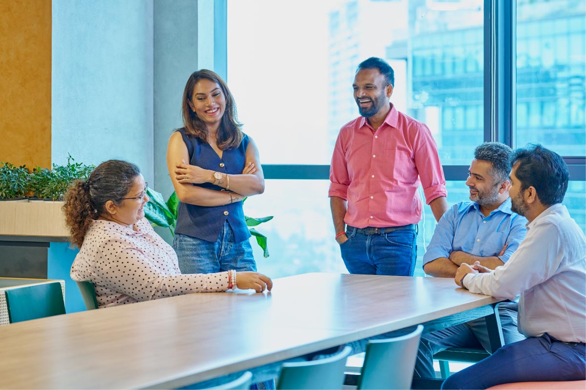 Group of colleagues standing and sitting around a meeting table, smiling and having a relaxed conversation in a bright office.