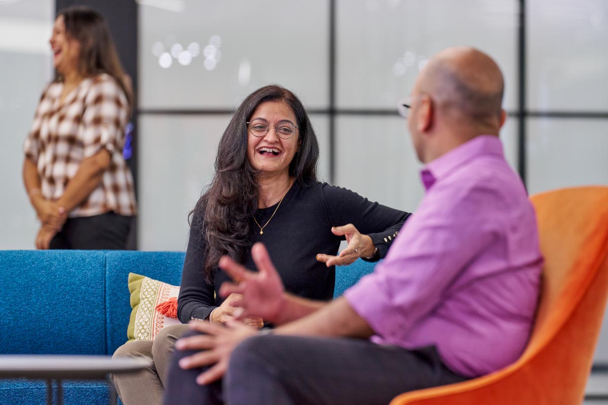 Smiling woman in conversation with a colleague, gesturing as she speaks in a bright office setting.