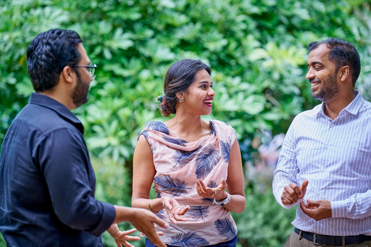 Two colleagues smiling and talking outdoors with greenery in the background.