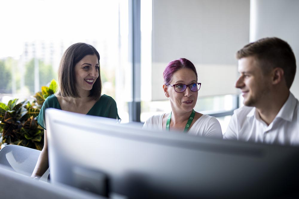 Three colleagues collaborating at a desk, smiling and talking while working at a computer in a bright office.
