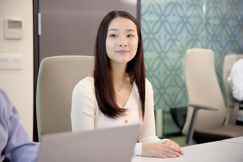 Young woman with long straight hair sitting in a modern office chair, looking thoughtful with a slight smile in a meeting room.