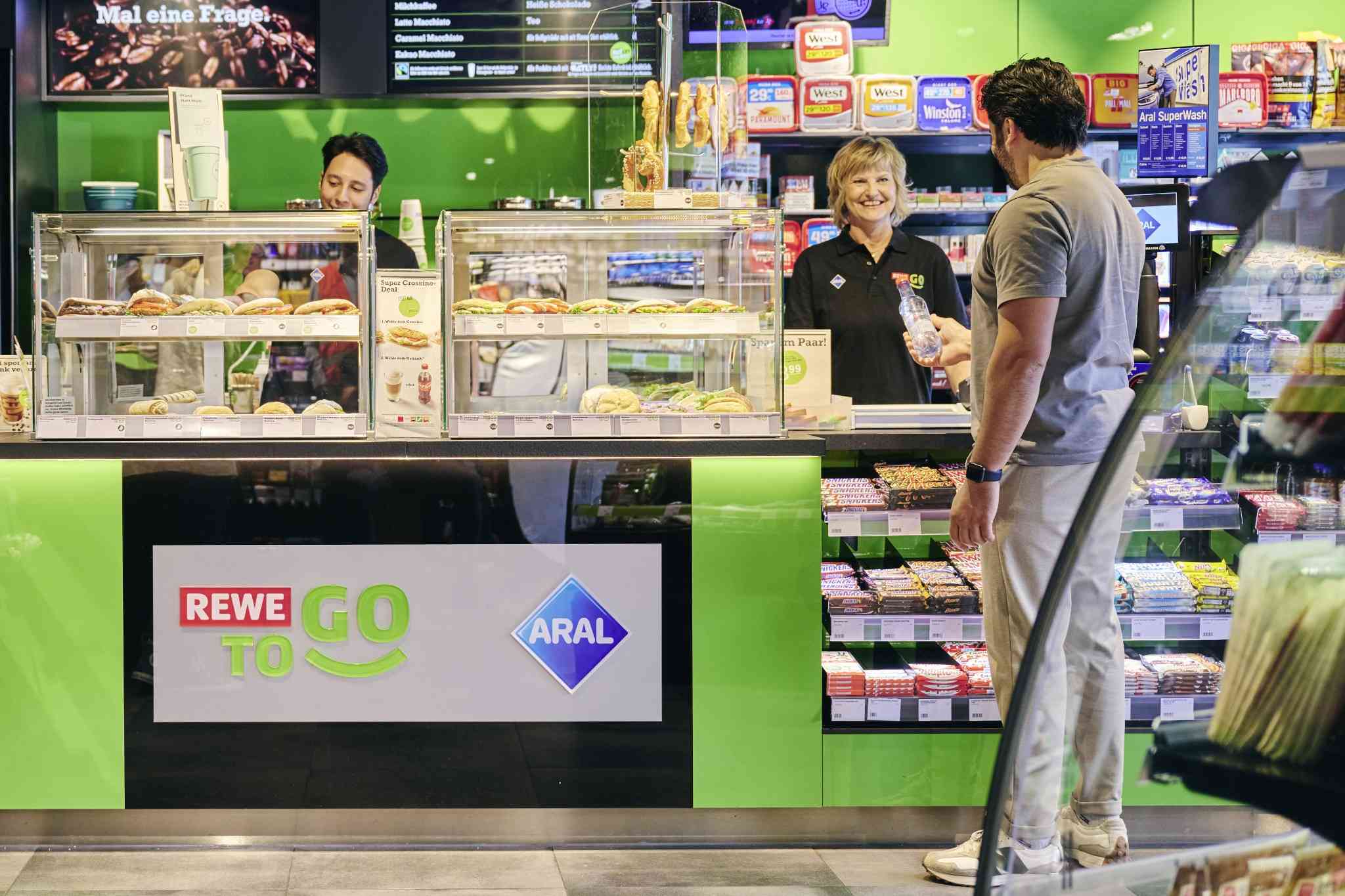 Customer ordering food at a REWE To Go counter inside a convenience store.