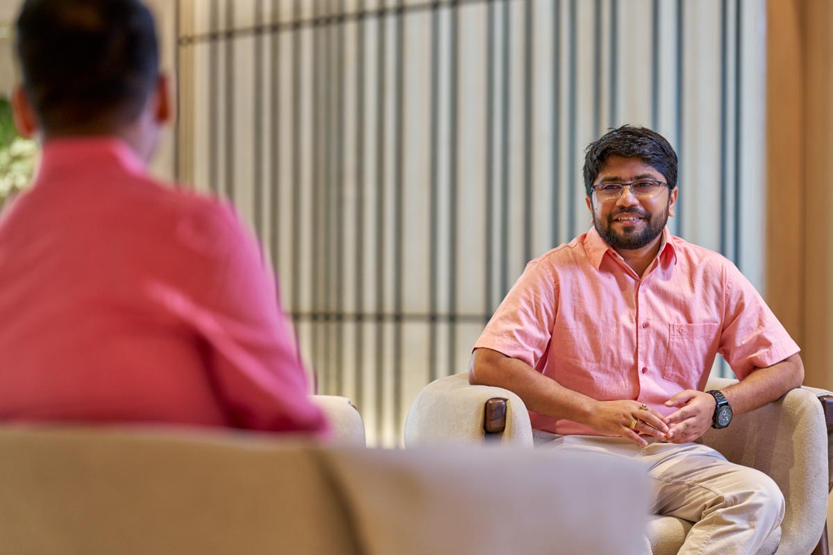 Two colleagues in conversation, one smiling while seated and facing another person in an office setting.