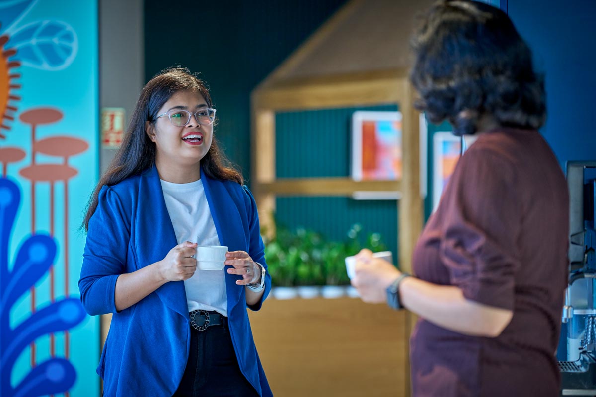 Two colleagues talking in a brightly colored office setting, with one person smiling during the conversation.