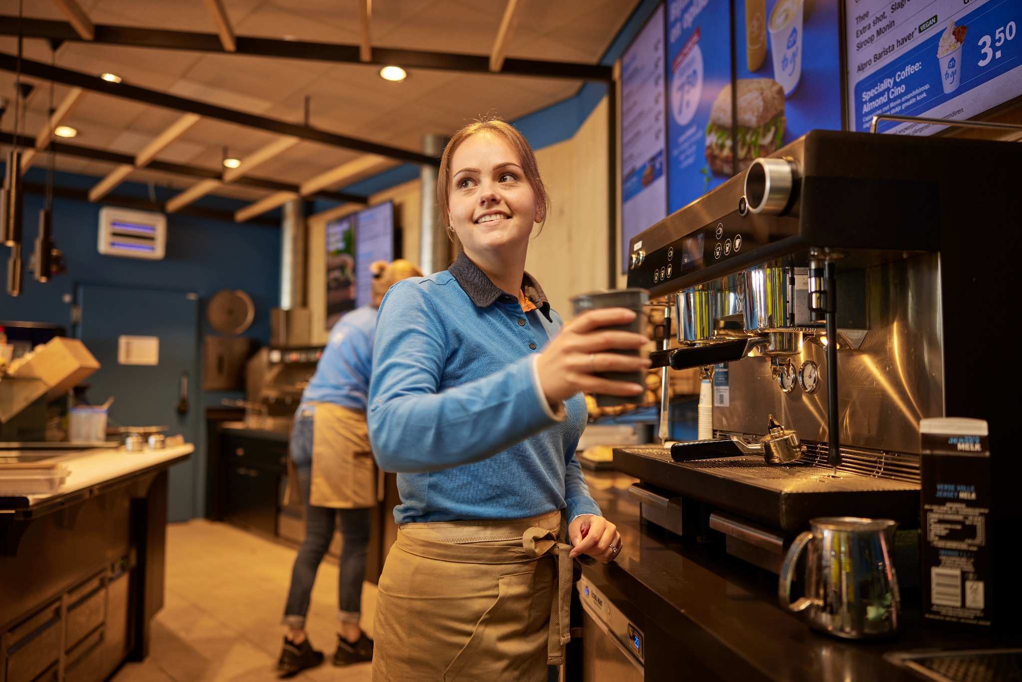 Barista in blue uniform serving coffee at an espresso machine.