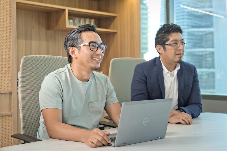 Two men sitting side by side in a modern office, one smiling in a casual t-shirt, the other in a blazer and glasses, both engaged in a meeting.