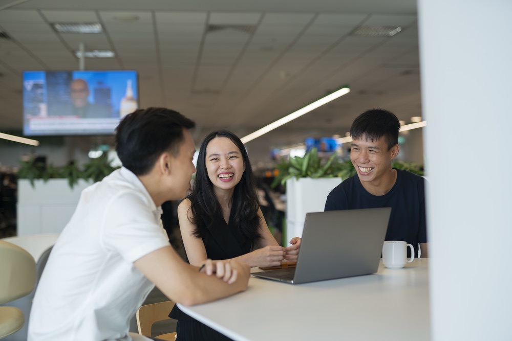 Three colleagues smiling and talking around a laptop in a modern office.