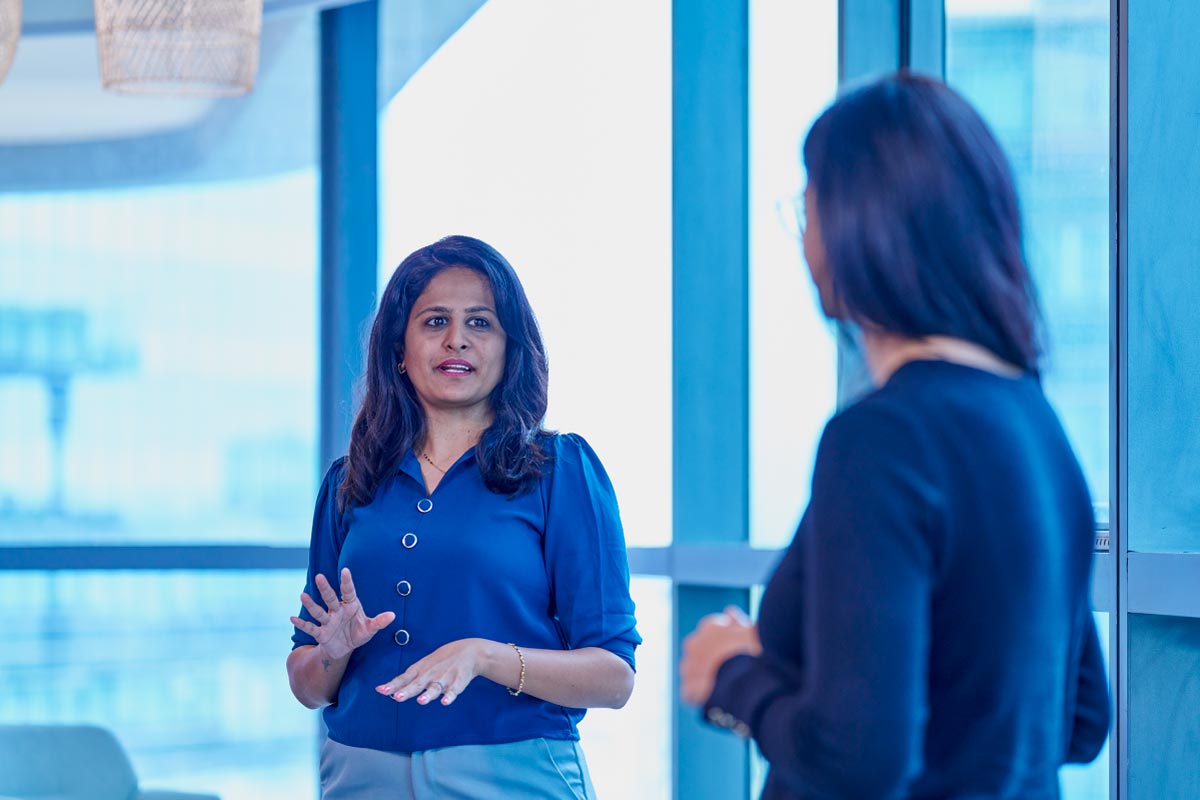 Woman in a blue blouse speaking to a colleague in an office with large windows.