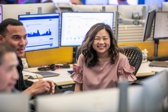 Team of colleagues smiling and talking while working at computer desks with monitors displaying data