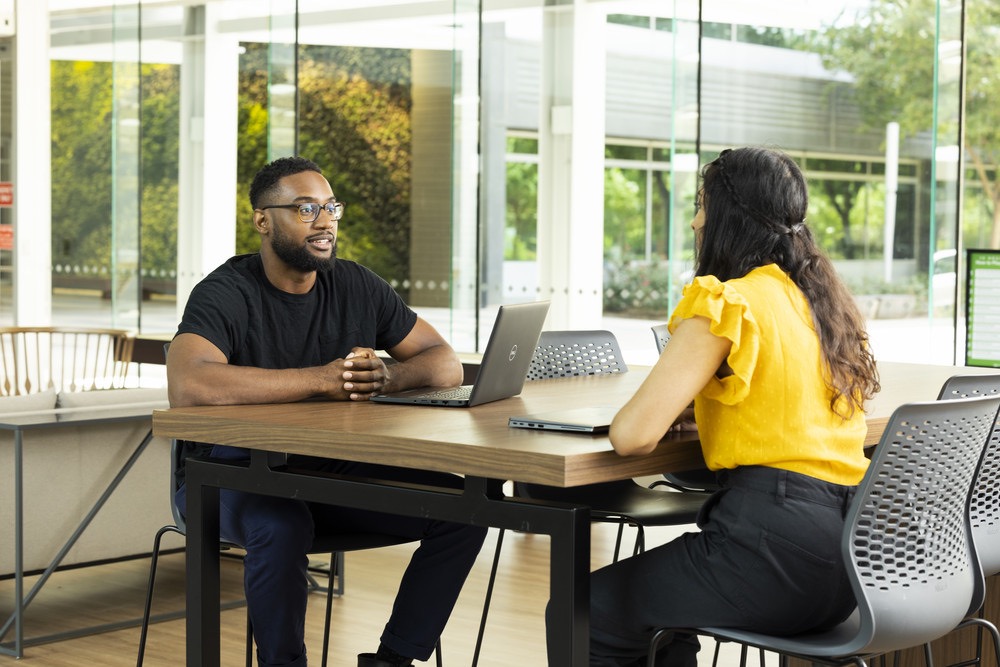 Two colleagues talking at a table with laptops in a modern office.