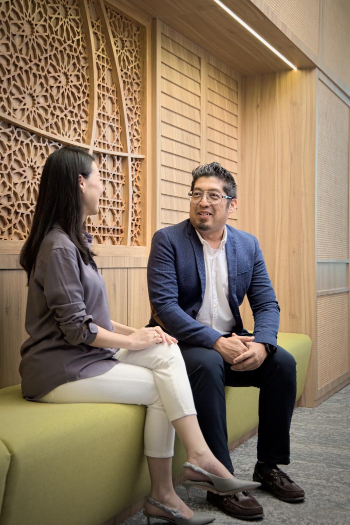 Two people sitting indoors having a conversation. A man in a suit jacket and glasses is speaking while a woman listens and smiles.