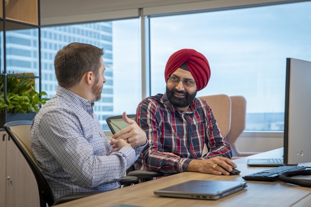 Two bp employees in an office having a discussion. One man in a checked shirt and red turban is smiling while listening to a colleague.