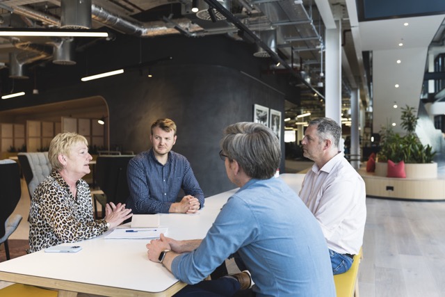 Four people seated around a white table in a modern office space, engaged in a group discussion.
