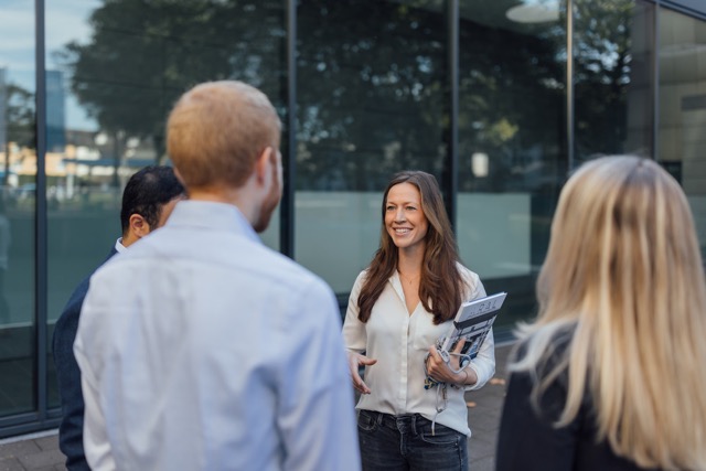 Female employee smiling and speaking with colleagues outdoors during a team meeting.