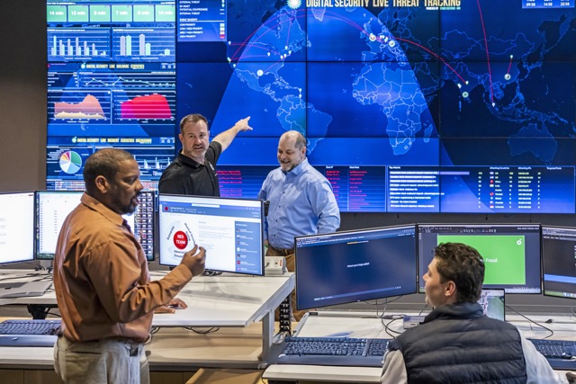 Team of employees in a security operations center reviewing data dashboards and world maps on large screens.