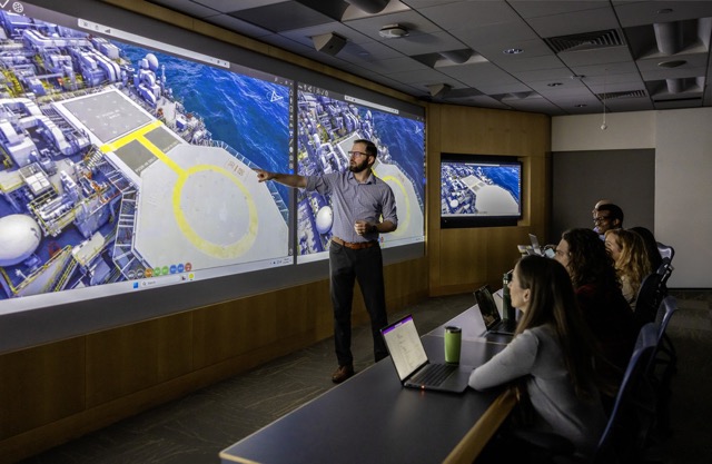 Employee giving a presentation with 3D visuals of an offshore facility projected on large screens.