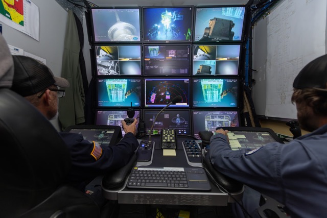Two operators controlling underwater equipment from a monitoring station with multiple screens.