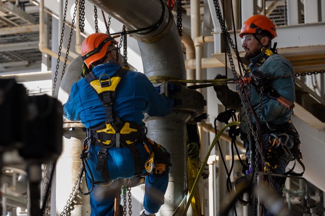 Two oil and gas workers wearing safety harnesses and helmets securing large industrial pipeline with chains.