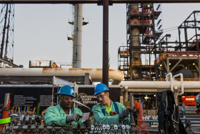 Two industrial workers in safety helmets and green protective uniforms inspecting equipment at oil refinery site.
