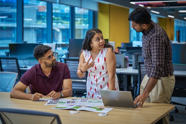 Team collaboration in modern office setting with three colleagues discussing ideas at their workspace.
