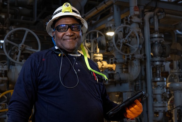 Oil and gas worker smiling in protective gear and helmet with industrial pipes in the background.