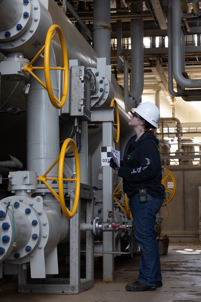 Female engineer in safety helmet inspecting industrial equipment and pipelines at an energy facility.