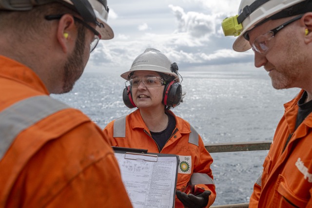 Three offshore oil and gas workers in safety helmets and protective gear having a discussion at sea platform.