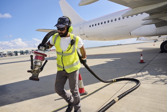 Airport ground worker in high-visibility vest handling refueling hose beside an airplane.