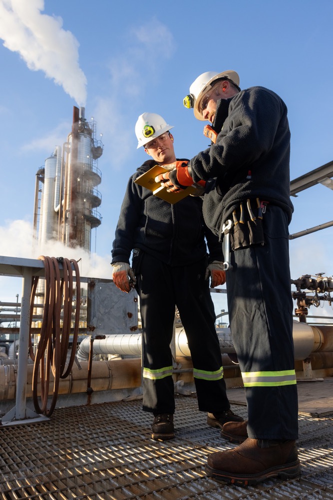 Industrial workers in hard hats reviewing notes at oil refinery with storage towers and steam in background.