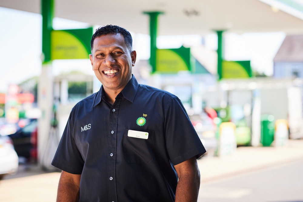 bp service station employee smiling in front of fuel pumps, representing customer service and sustainable mobility.