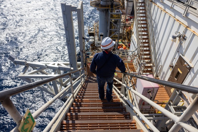 Employee in safety gear walking down stairs on an offshore platform above the ocean.
