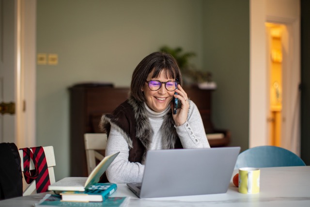 Woman working from home, smiling while talking on the phone and using a laptop at a table.