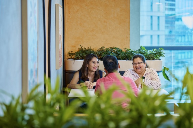 Three people sitting together at a table in a bright office space, smiling during a conversation.