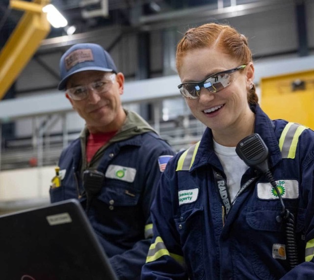 Two bp industrial workers in protective gear smiling while looking at a laptop in a facility.