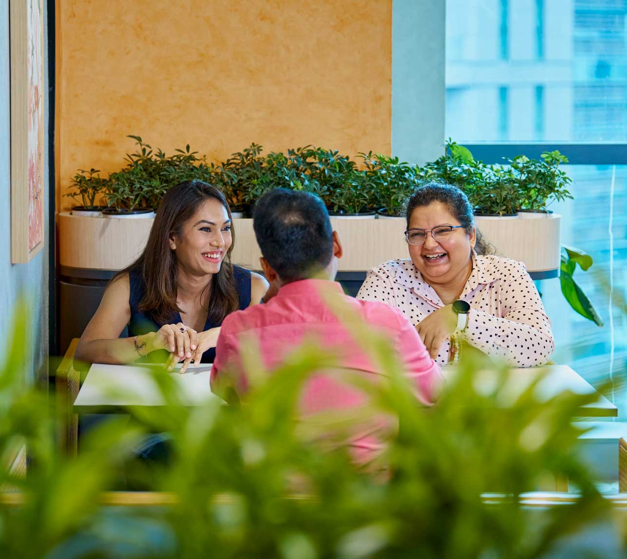 Three colleagues sitting at a table, smiling and talking in a bright office space.