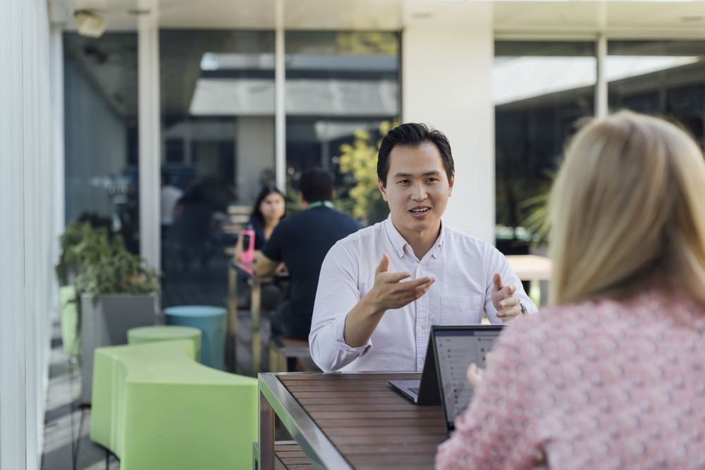 Two people sitting outdoors at a table with a laptop, engaged in a conversation, while others are seated in the background.
