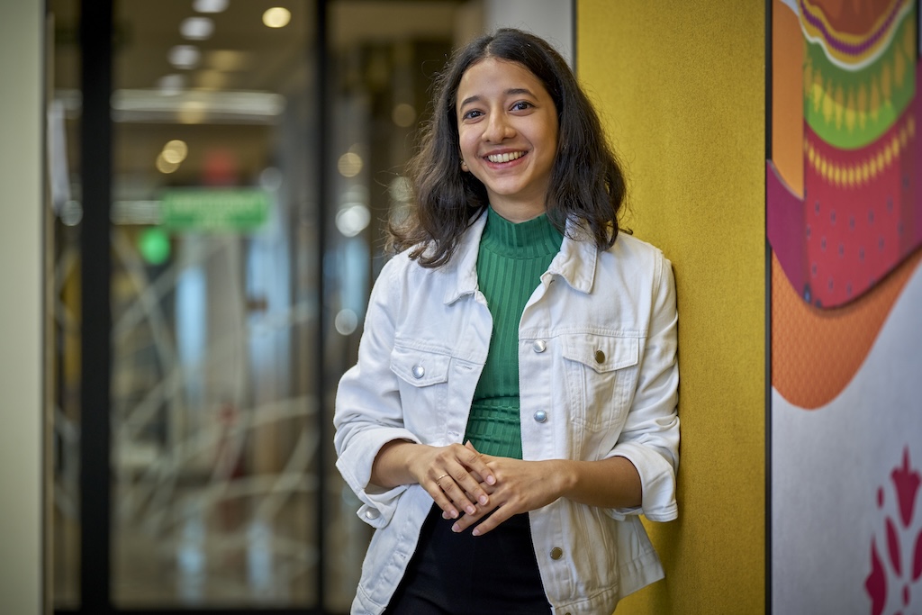 Woman standing indoors, smiling and leaning against a yellow wall with colorful artwork.