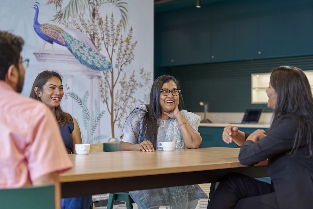 Group of colleagues smiling and talking over coffee at workplace table.
